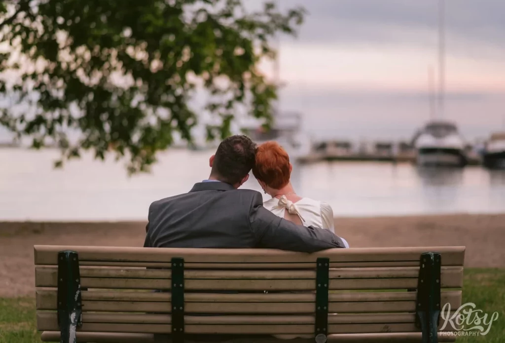 A bride and groom sit on a wooden park bench with their hands leaned into each other at a beach in Toronto