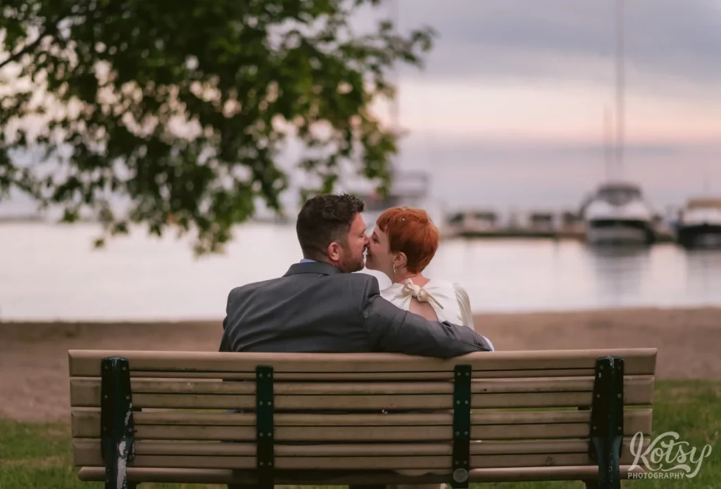 A bride and groom enjoy a kiss on a park bench at a beach in Toronto