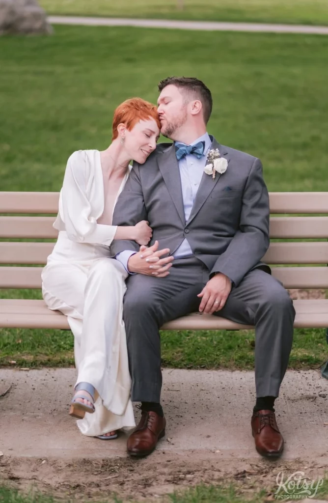 A bride smiles with her eyes closed at her groom kisses her on the forehead as they sit on a park bench in Toronto