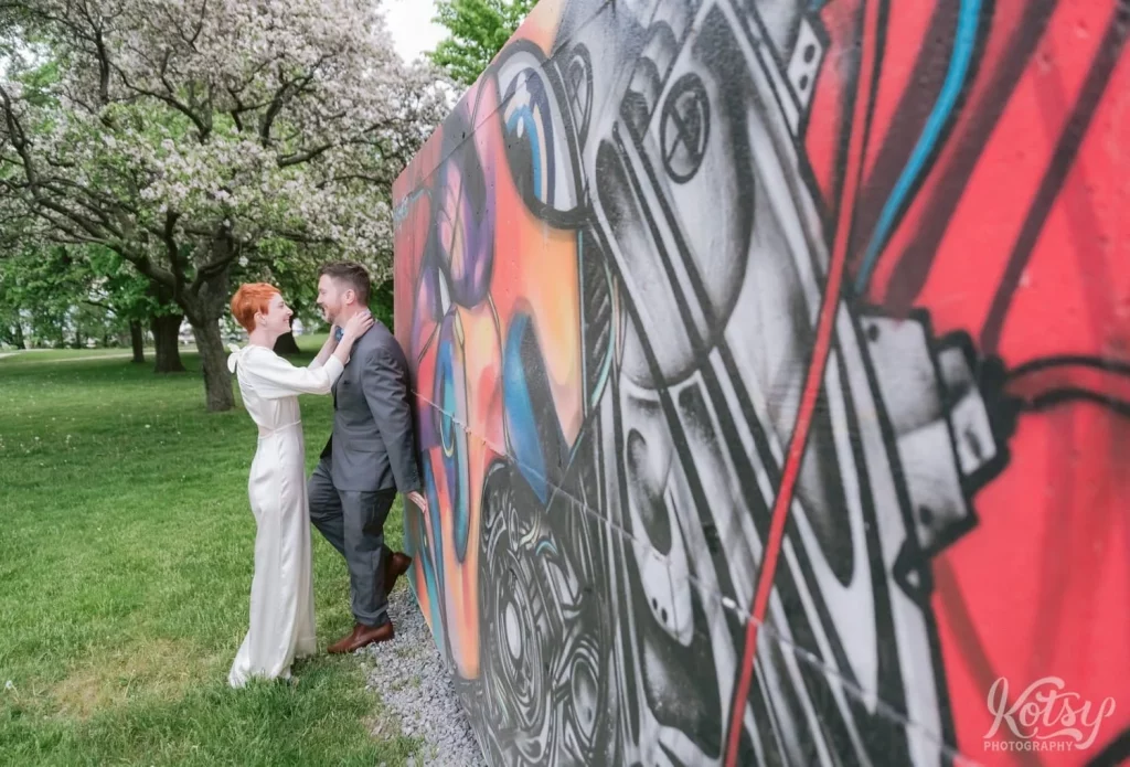 A bride smiles as she touches his neck while leaned up against a mural in a Toronto park