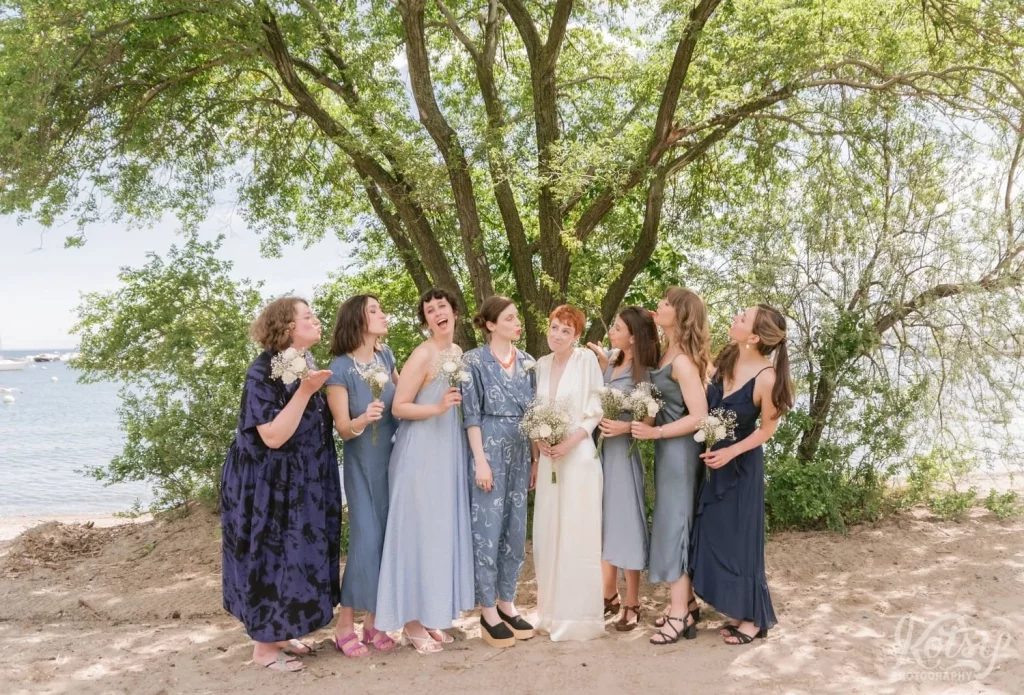 A bride and her bridesmaids enjoy a laugh in front of a large tree on a beach in Toronto