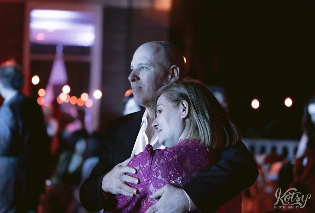 A man hugs his partner as they enjoy a band's performance at an outdoor wedding reception.