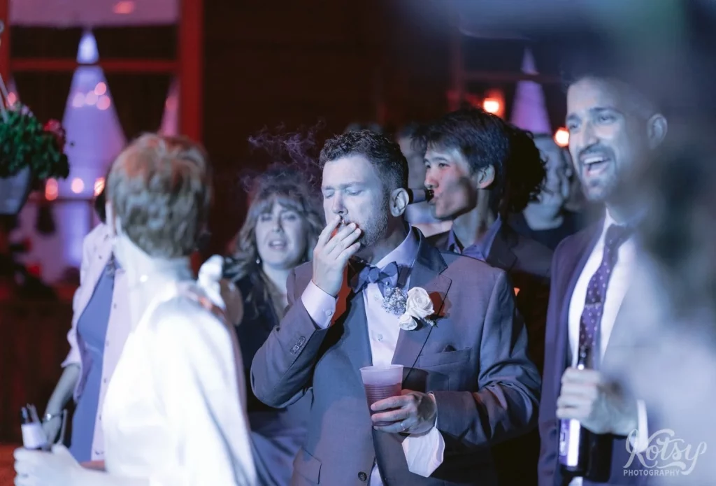 A groom puffs on a joint in a crowd of people during an outdoor wedding reception.