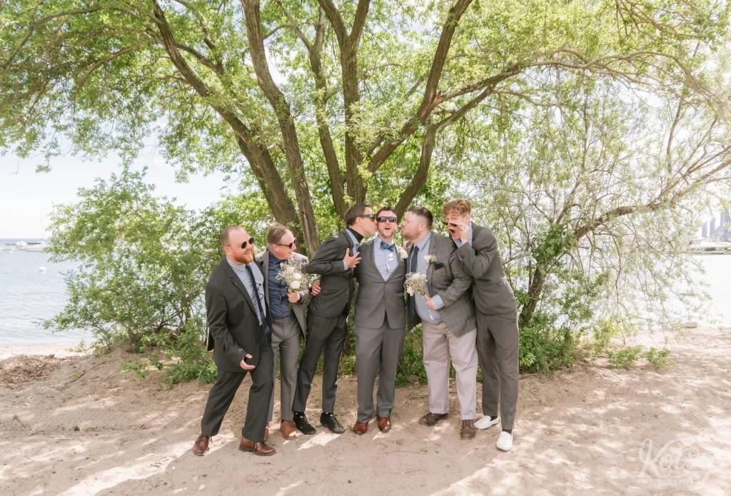 A groom goofs around with his groomsmen in front of a large tree on a beach in Toronto
