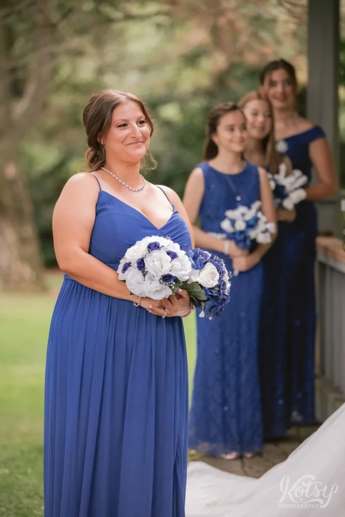 A maid of honour smiles while holding two flower bouquets. 3 young bridesmaids are seen in the background.