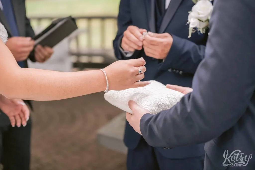 A close up of a bride's hands picking up a ring from a pillow during an outdoor wedding ceremony.