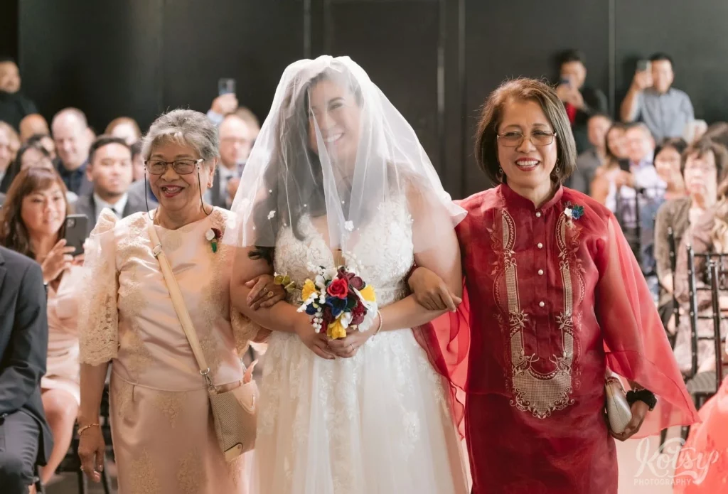 A bride smiles big as she approaches the end of the aisle with her mother and grandmother at The Vue Event Venue