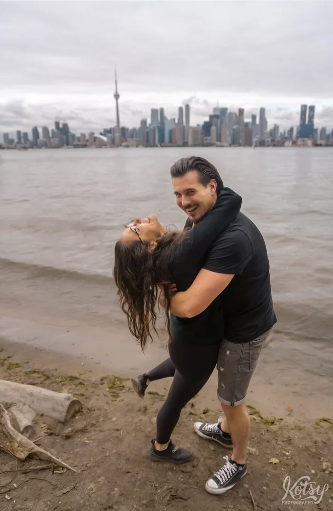 A man smiles largely at the camera as his new fiancé dips backwards out of shock after getting engaged at Toronto Islands