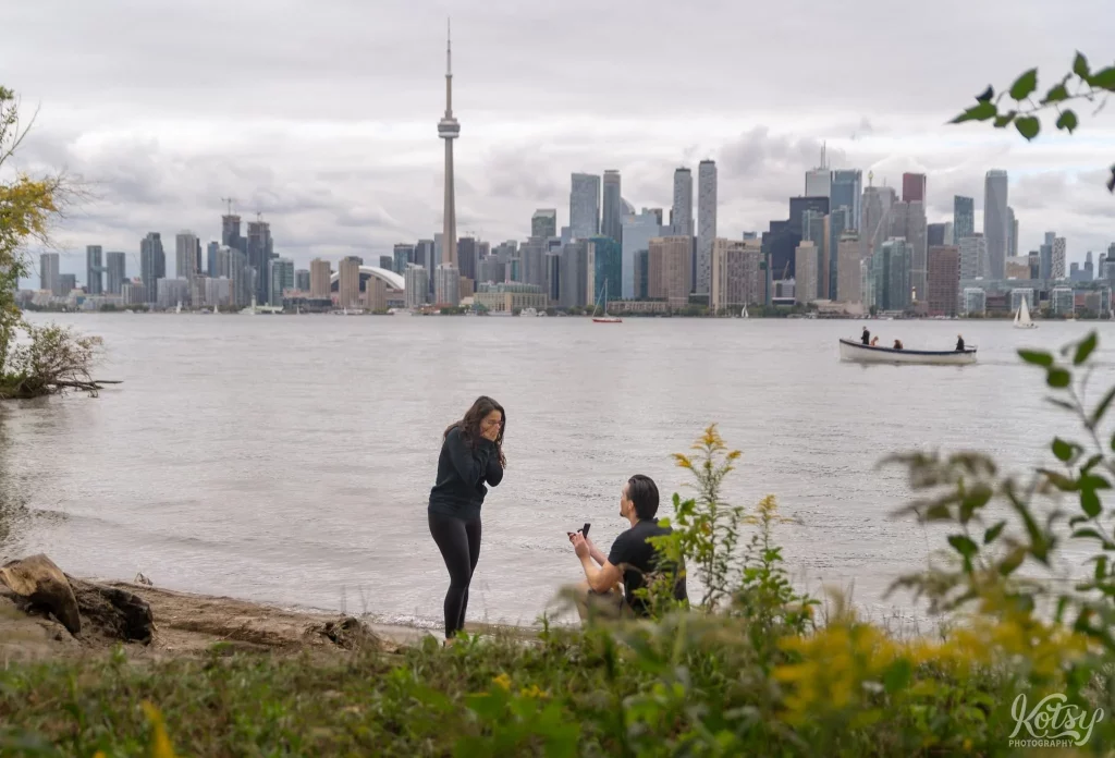 A man is down on one knee proposing to his girlfriend on a beach at Toronto Islands