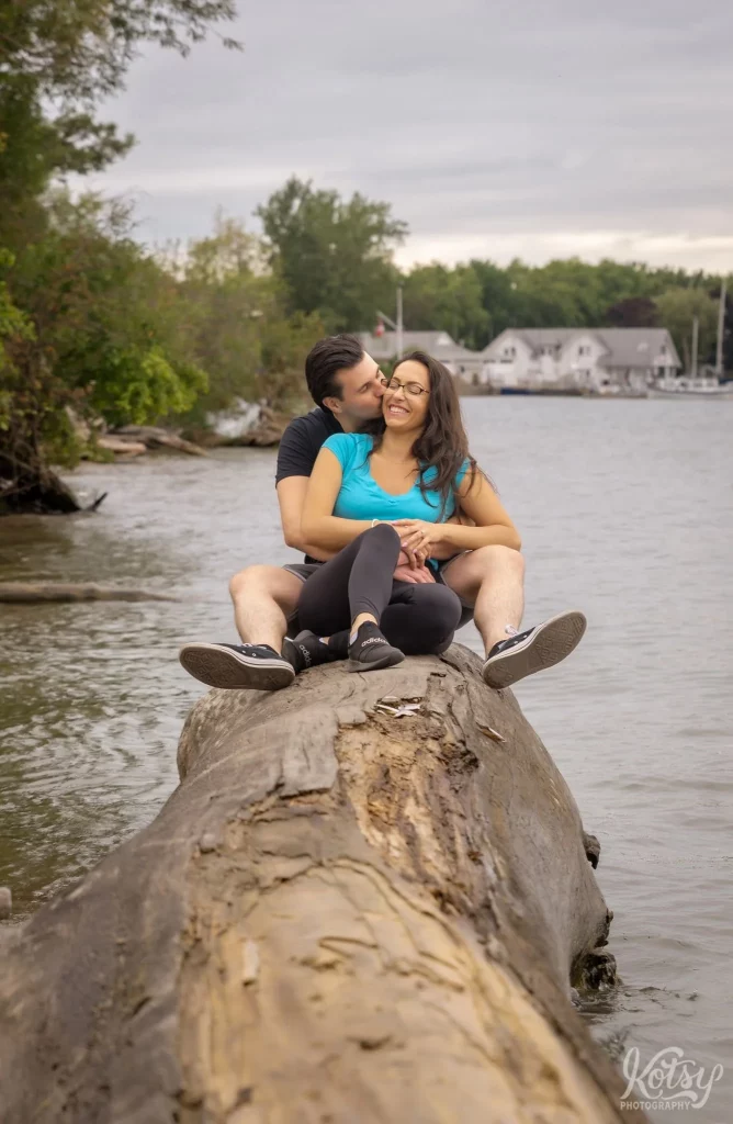 A man kisses his new fiancé on the cheek while sitting on a big log at Toronto Islands