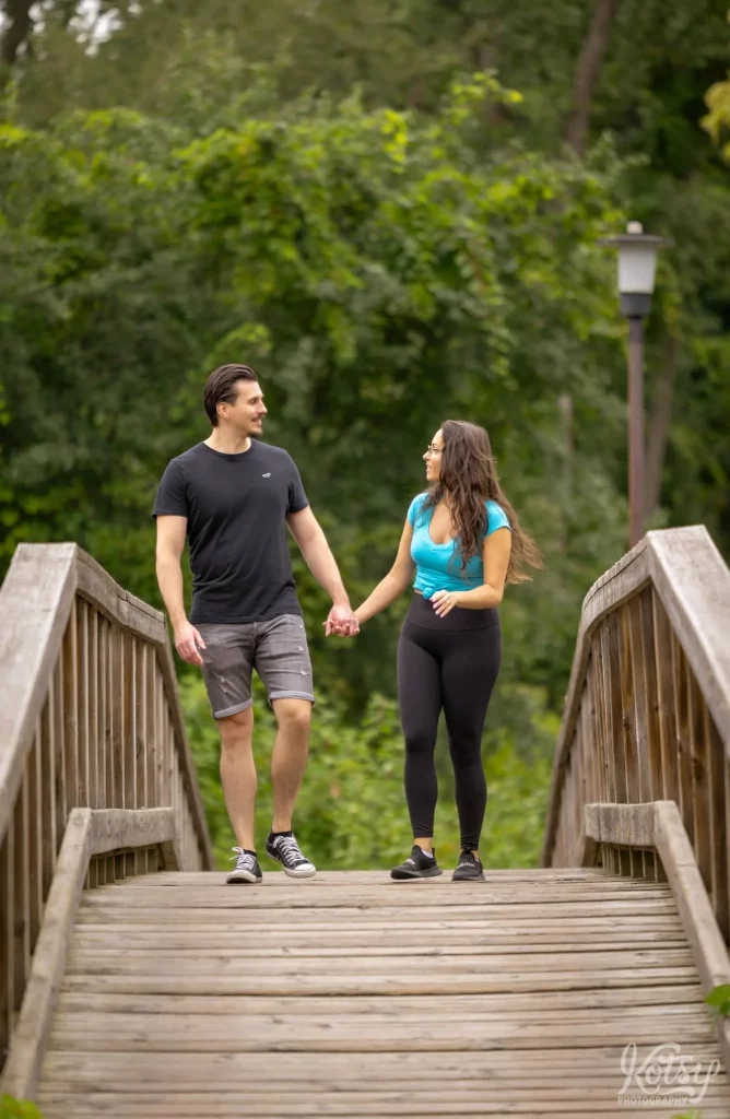A couple walk on a wooden bridge at Toronto Islands while smiling at each other