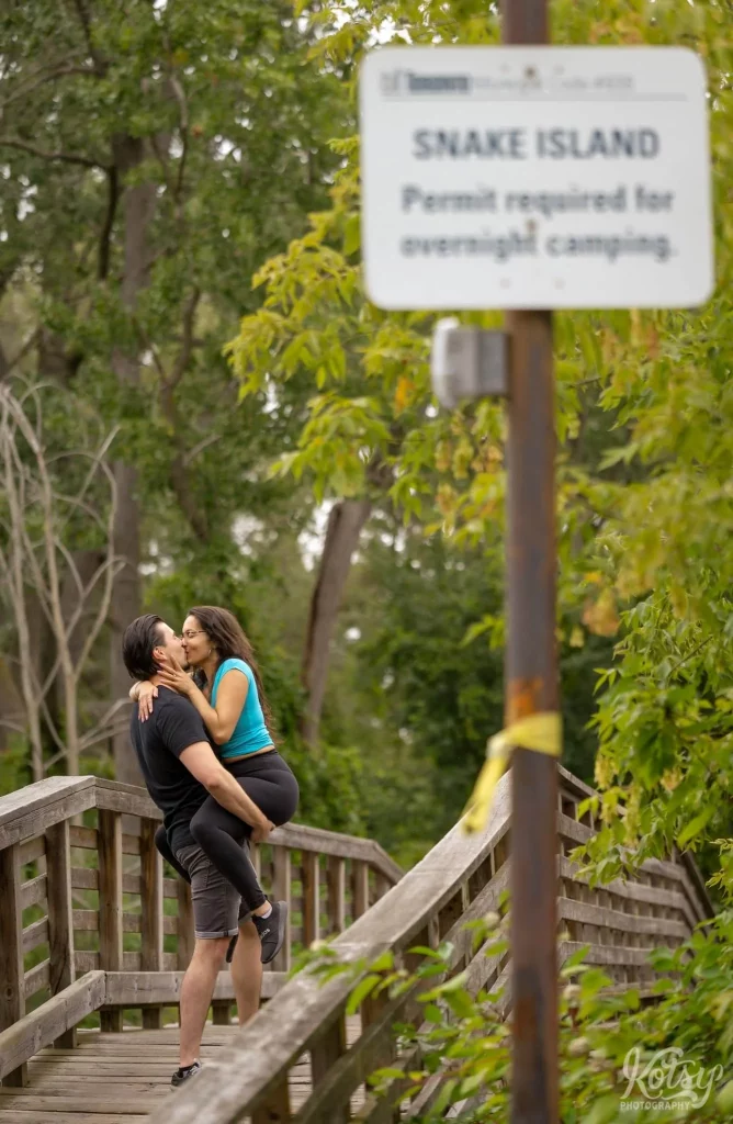 A woman kisses her fiancé holds her up on a wooden bridge on Snake Island in Toronto