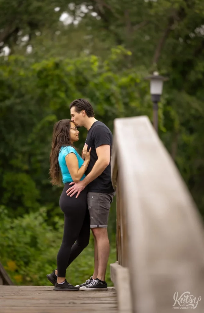 A man kisses his new fiancé on the forehead while standing on a wooden bridge