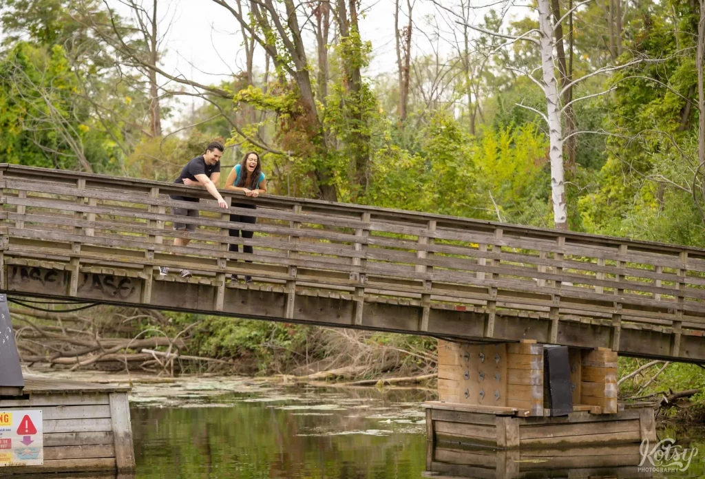 A man points to something in the water for his fiancé while on a wooden bridge