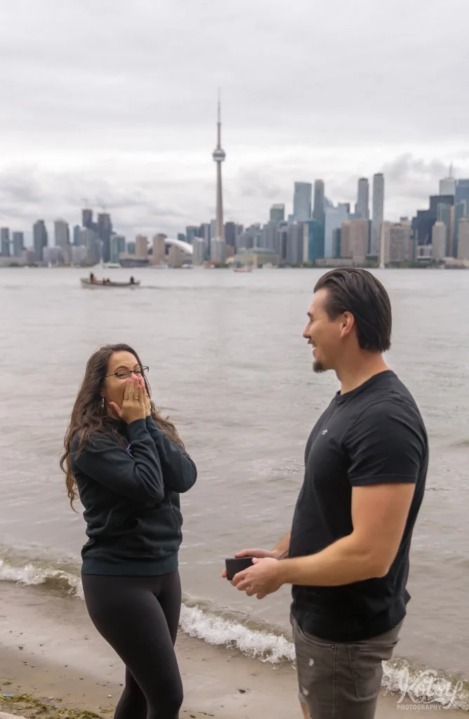 A woman covers her face in shock after her boyfriend proposes to her on a beach at Toronto Islands