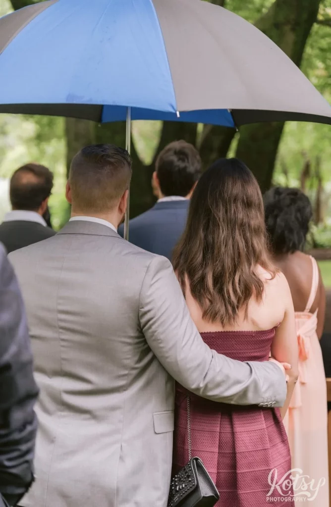 A man holding an umbrella and arm around his partner's waist watching a wedding ceremony in Edwards Gardens