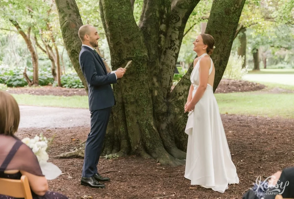 A groom reads vows to his bride with a big smile under a tree at Edwards Gardens in Toronto