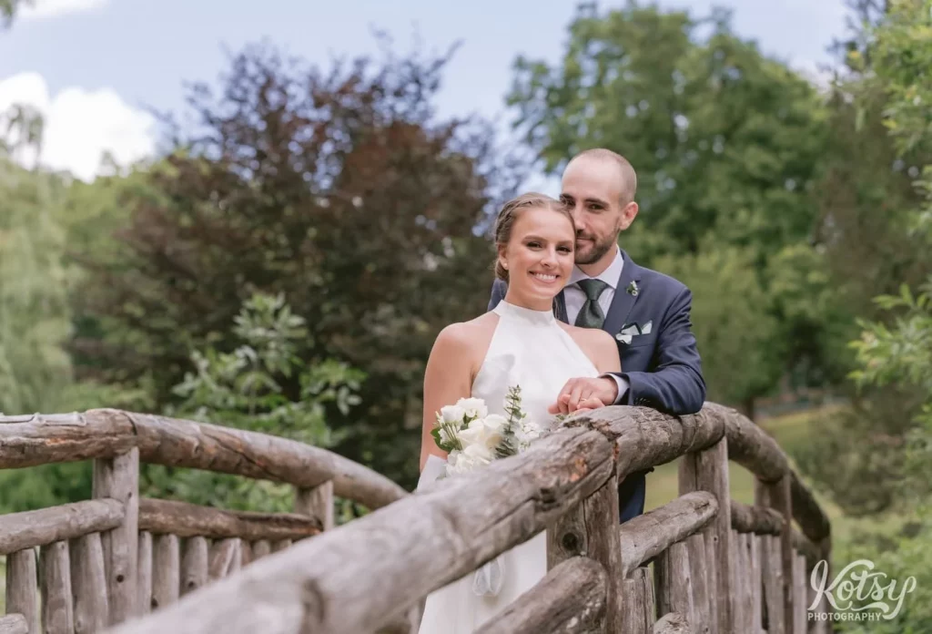 A bride and groom pose for a photo on a wooden bridge at Edwards Gardens in Toronto