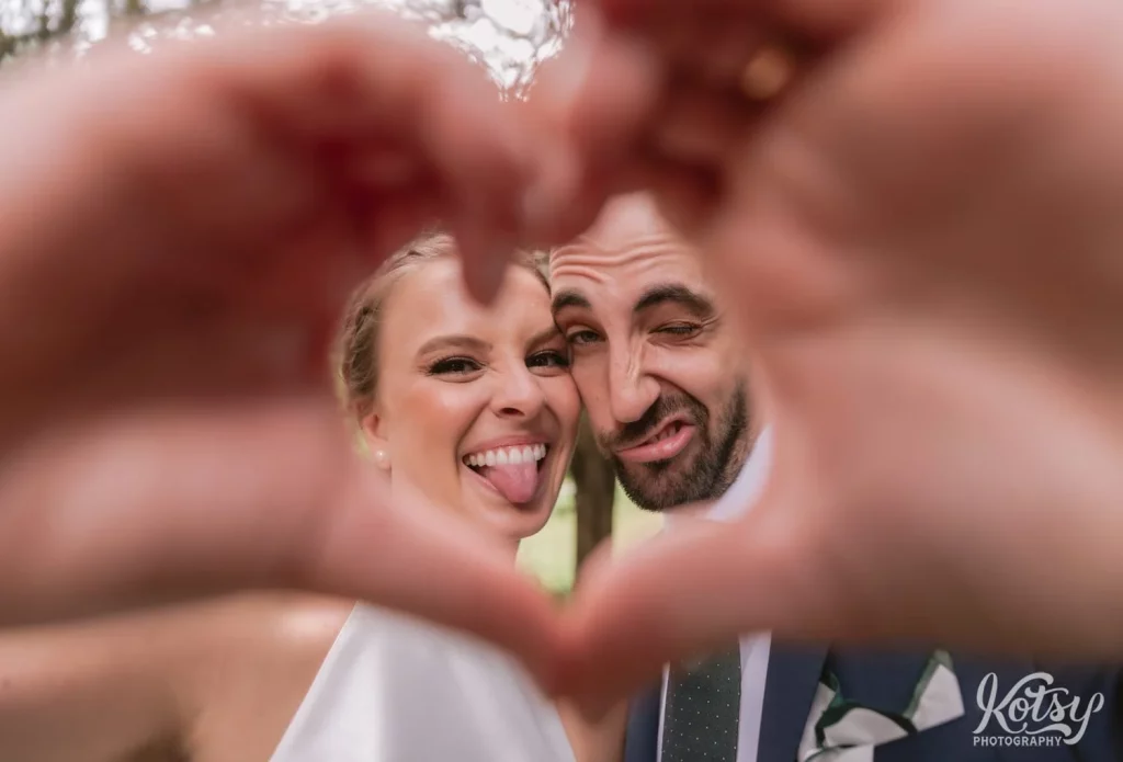 A bride and groom make funny faces behind their hands forming a heart for the camera.