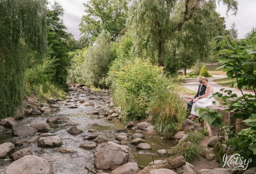 A bride and groom cozy up with each other while sitting on a big stone by a small river at Edwards Gardens