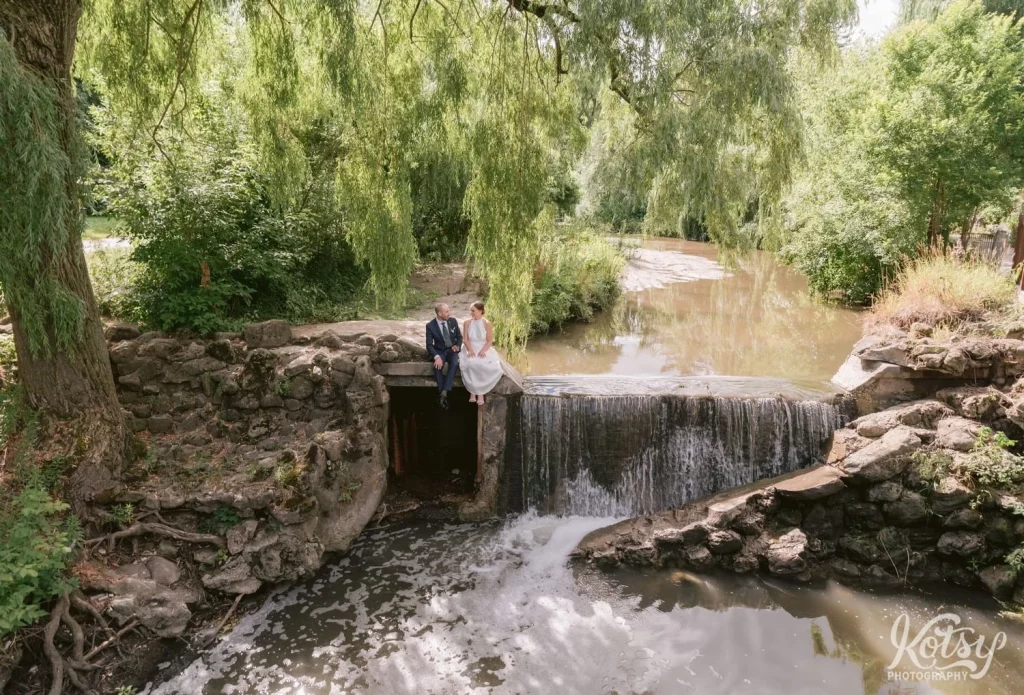 A wide shot of a bride and groom sitting next to a waterfall at Edwards Gardens in Toronto