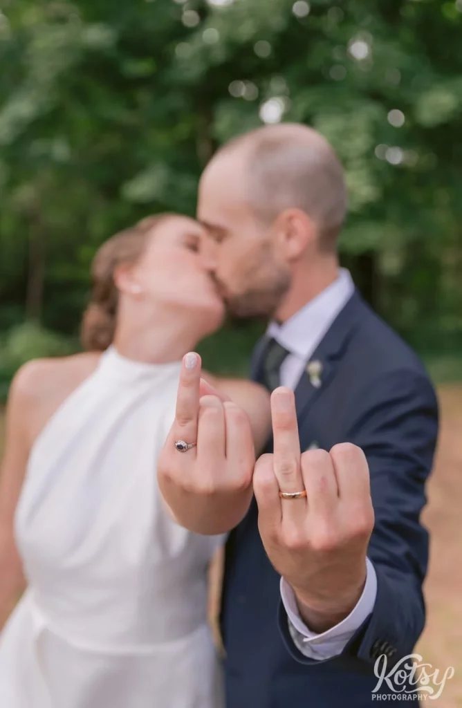 A bride and groom kiss as they extend their ring fingers at the camera