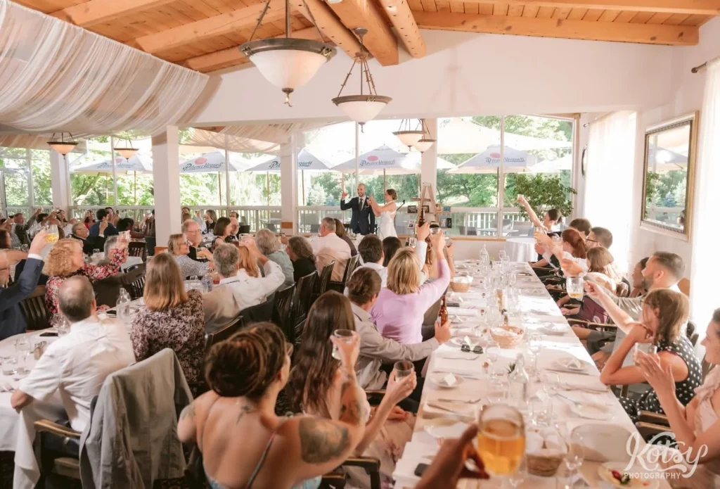 A room full of people raise their glasses to cheers the bride and groom during their wedding reception in Scarborough, Canada