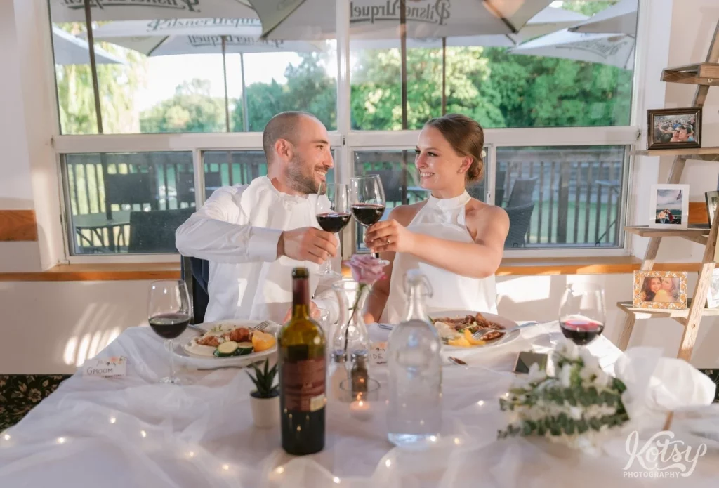 A bride and groom clink their wine glasses while eating dinner at their wedding reception at The Prague Restaurant in Scarborough
