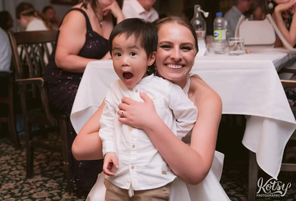 A bride smiles big while holding a child with his mouth wide open. Photographed at her wedding reception at The Prague Restaurant in Scarborough