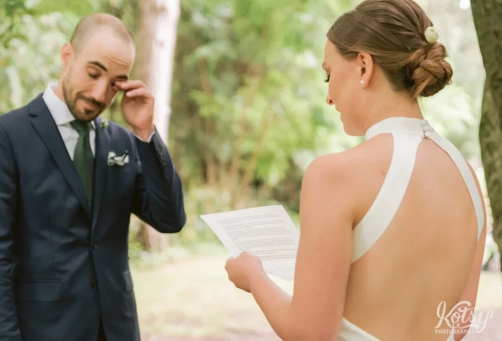 A groom wipes a tear from his eye as his bride reads her wedding vows in Edwards Gardens