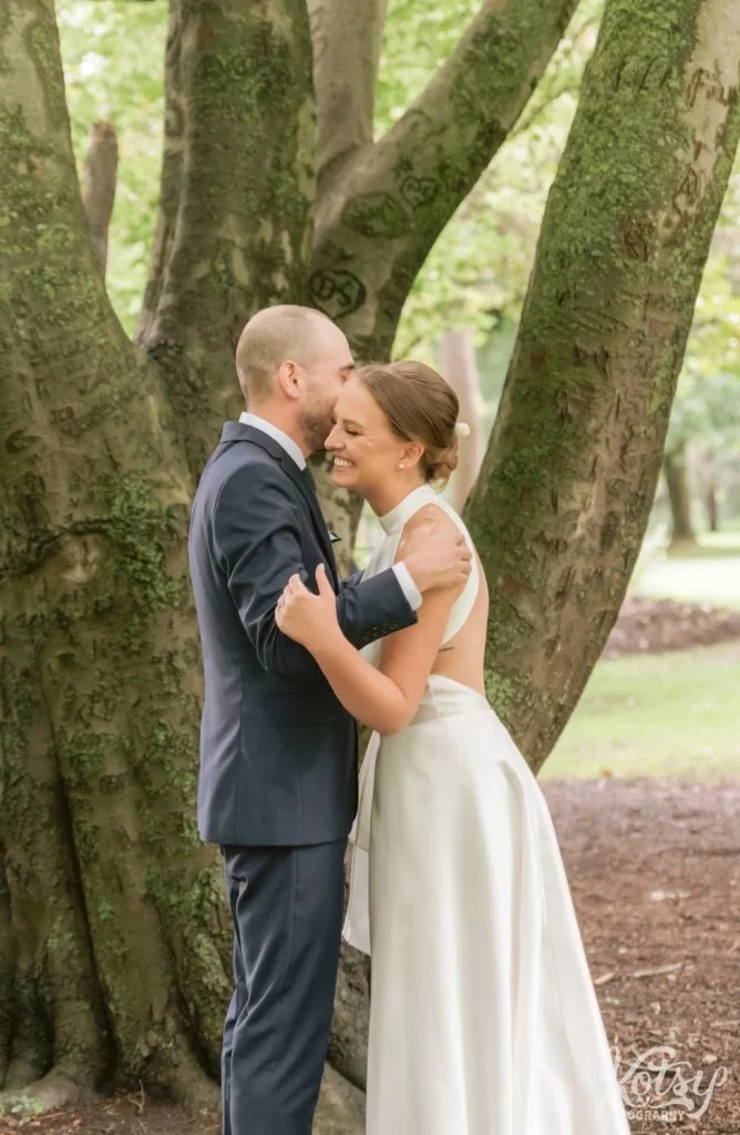 A bride smiles big as her groom whispers into her ear under a big tree at Edwards Gardens in Scarborough