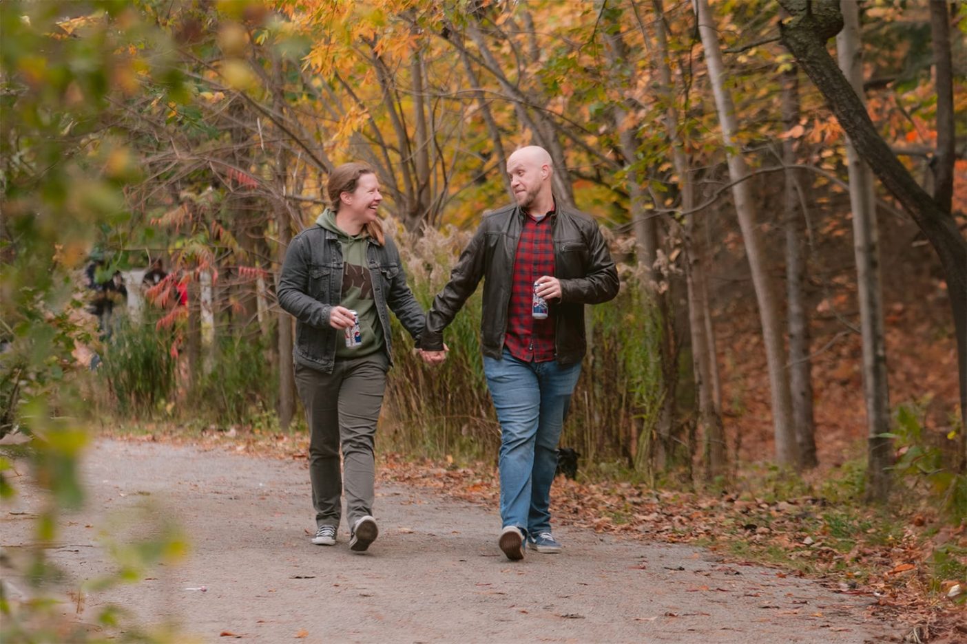 An engaged couple with beers in hand go for a walk through a forest during fall