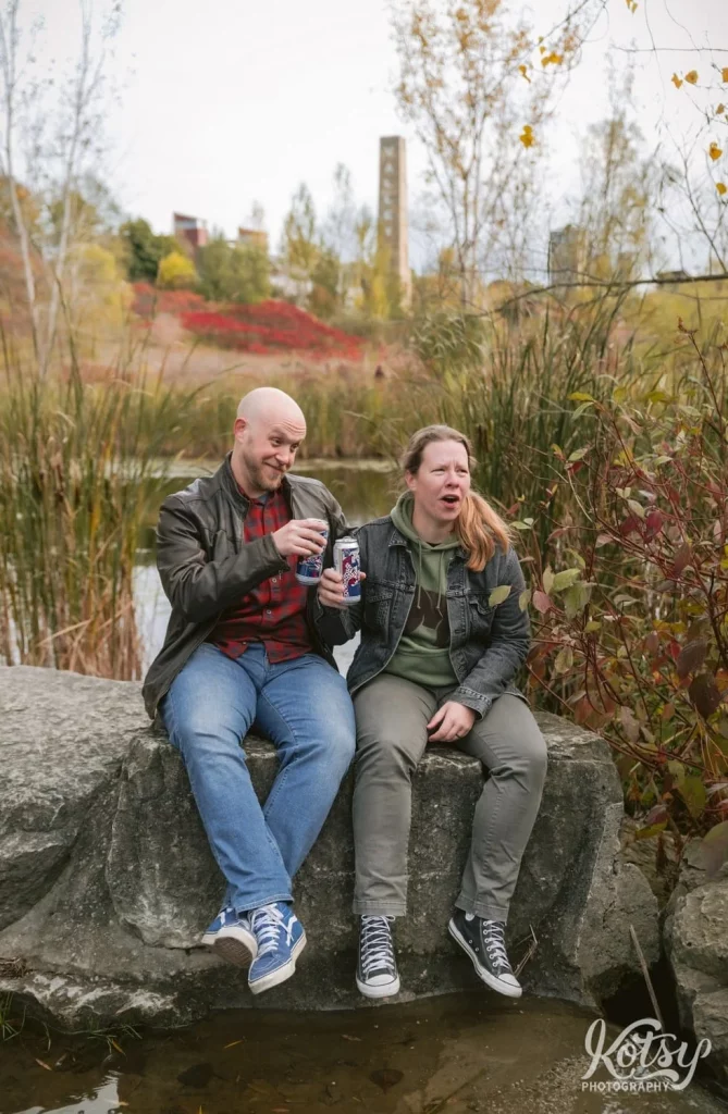 A couple react to something off camera while sitting on a big stone amongst fall colours in Don Valley Brick Works Park in Toronto.