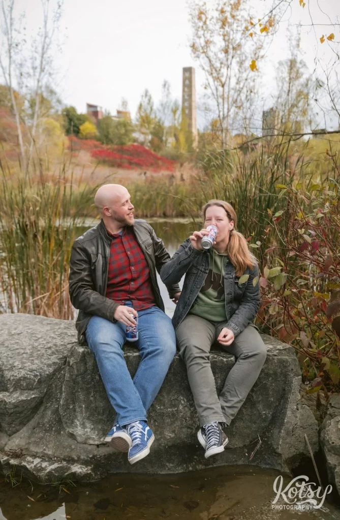 A man smiles at his partner as she sips on a beer while sitting on a big stone amongst fall colours in Don Valley Brick Works Park in Toronto.