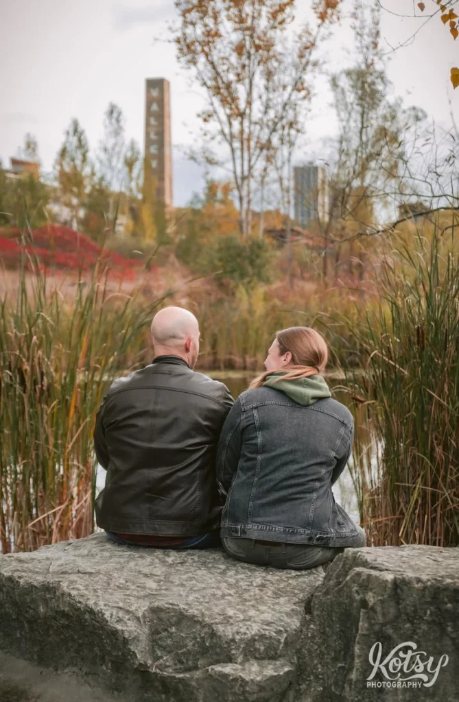 A shot from behind of a couple sitting on a big stone amongst fall colours in Don Valley Brick Works Park in Toronto.