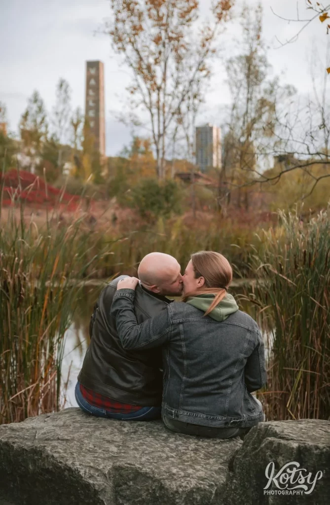 A shot from behind of a couple kissing while sitting on a big stone amongst fall colours in Don Valley Brick Works Park in Toronto.