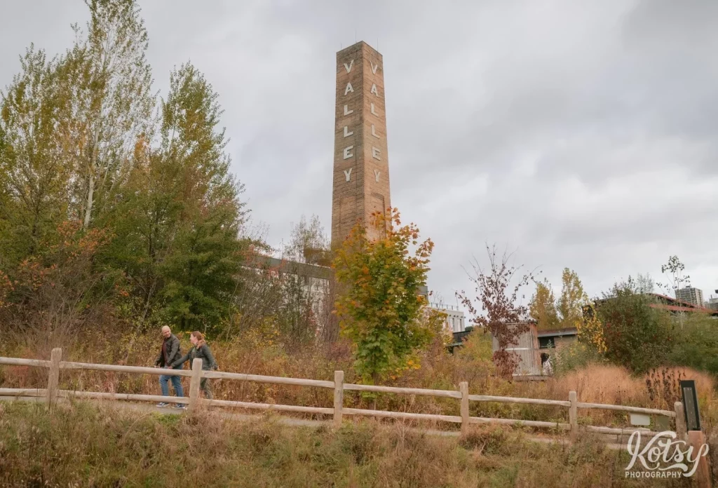 A couple walk-in-hand past a big chimey amongst fall colours in Don Valley Brick Works Park in Toronto.