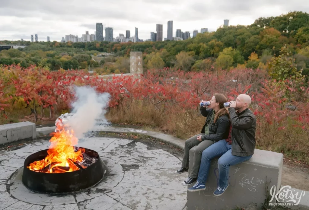 An engaged couple sip on beers near a camp fire at Governor's Bridge Lookout amongst fall colours in Don Valley Brick Works Park in Toronto.