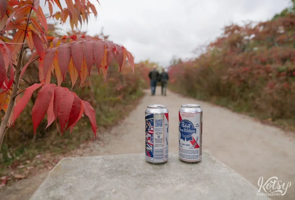 A tight focus on two cans of Pabst Blue Ribbon as an out of focus couple walks on a trail amongst fall colours in Don Valley Brick Works Park in Toronto