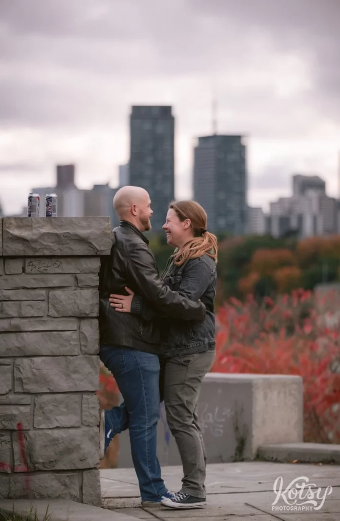 A couple have big smiles with their arms around each other's waist. Photographed on Governor's Bridge Lookout at Don Valley Brick Works Park in Toronto