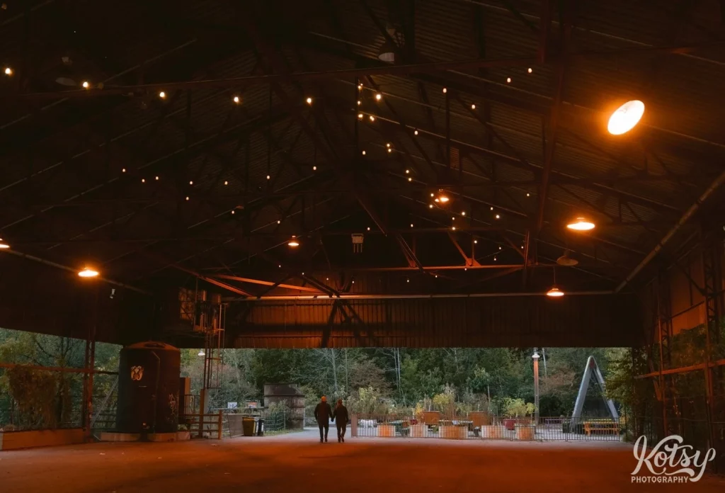 A really wide shot shows the silhouette of a couple walking away from camera in a large building with string lights over head and park behind them at Evergreen Bricks in Toronto