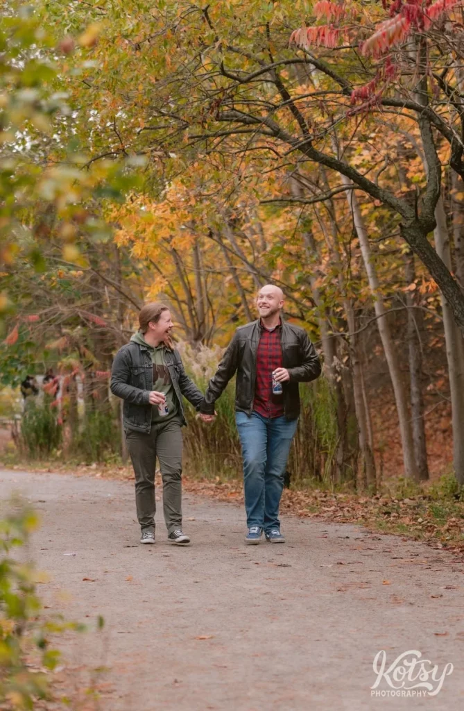 An engaged couple laugh as they walk with beers on a trail amongst fall colours in Don Valley Brick Works Park in Toronto.