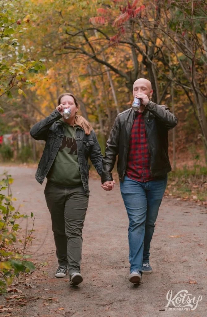 An engaged couple sip beers as they walk on a trail amongst fall colours in Don Valley Brick Works Park in Toronto.