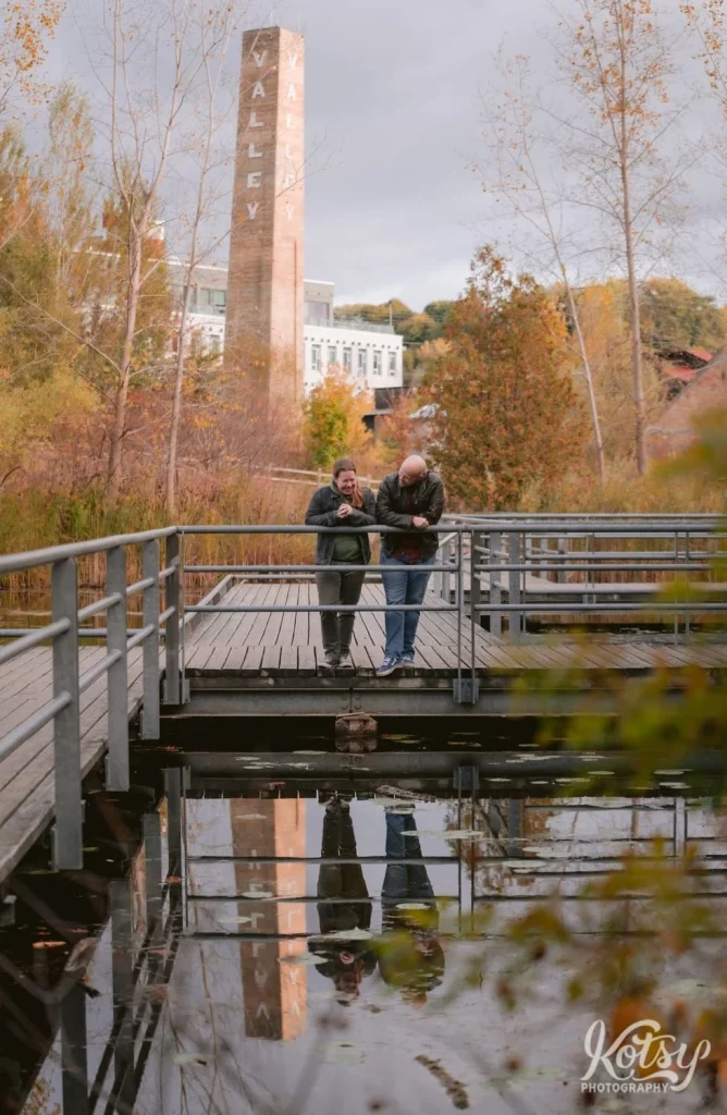 A couple stand with their arms leaning on a hand rail on a wooden dock surrounded by water and fall colours in Don Valley Brick Works Park in Toronto.