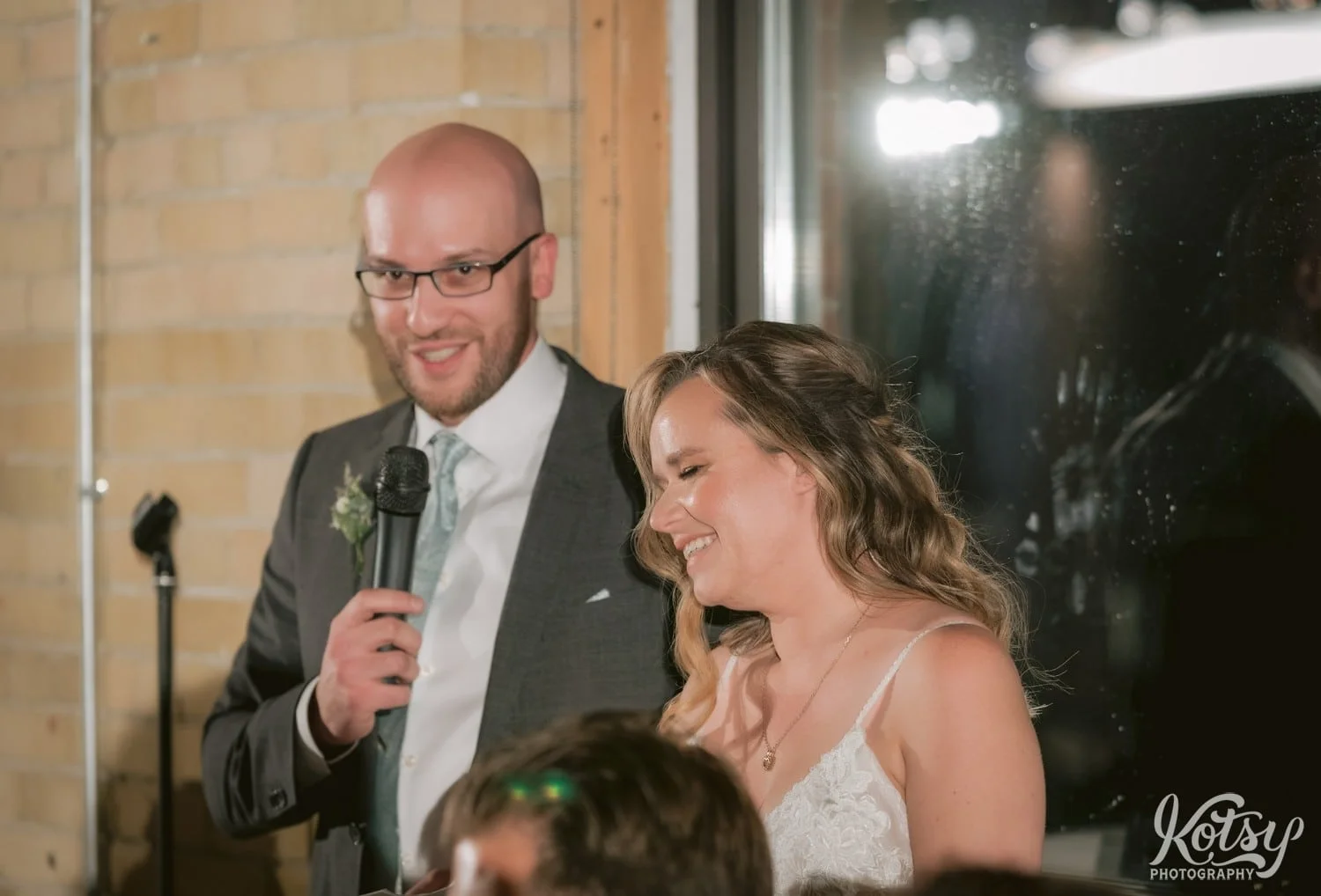 A bride in a white wedding gown enjoys a laugh as her groom in a gray suit and green tie speaks into a microphone during their Second Floor Events wedding reception in Toronto, Canada.