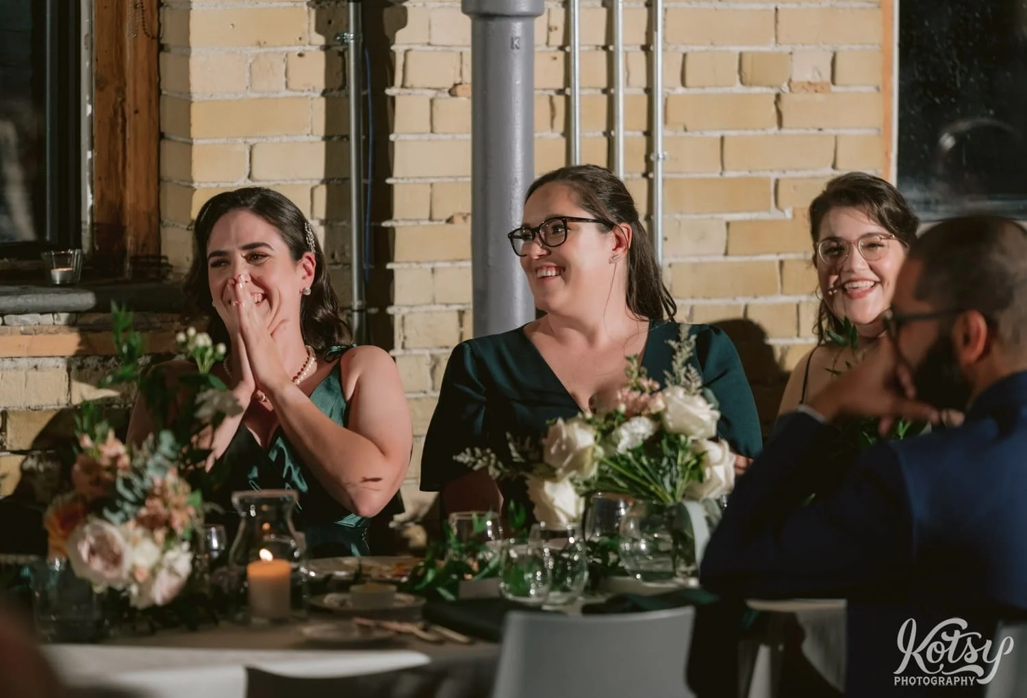 Three women in green dresses sitting down at a dinner table smile big in reaction to someone speaking off camera at a Second Floor Events wedding reception in Toronto, Canada.