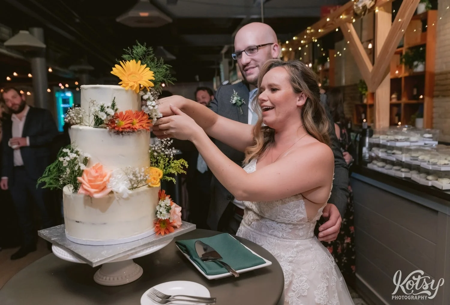 A bride in a white wedding gown and groom in gray suit cut a three tier wedding cake covered in flowers and white icing during their Second Floor Events wedding reception in Toronto, Canada.