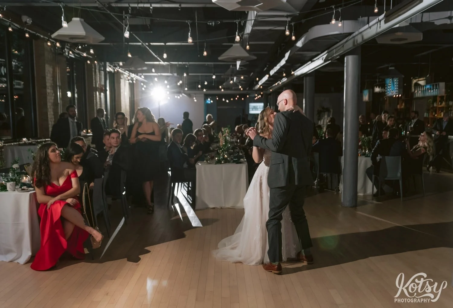 A bride in a white wedding gown and grooming gray suit enjoy a first dance in front of a large crowd of people sat at dinner tables during their Second Floor Events wedding reception in Toronto, Canada.