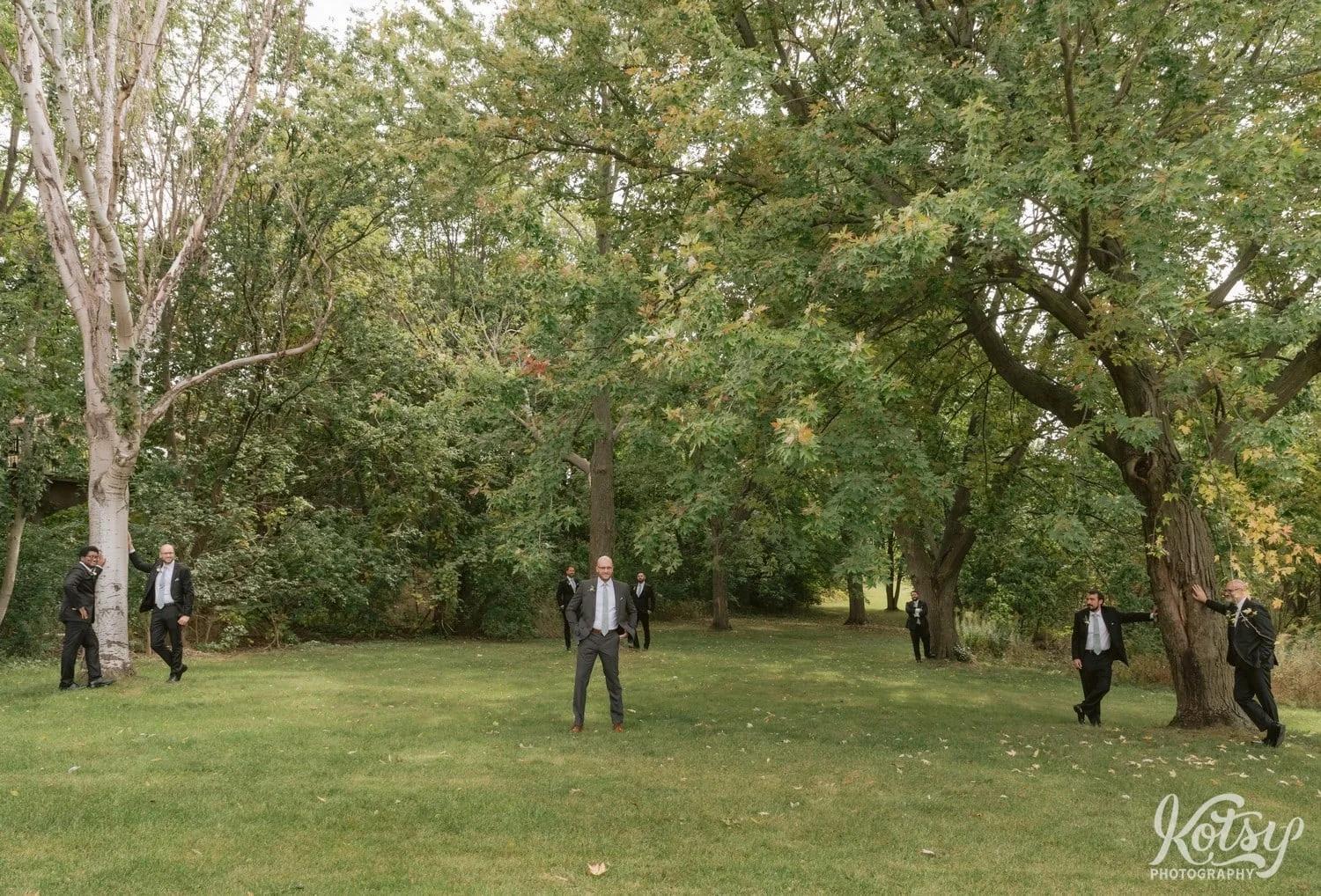 A groom and 7 groomsmen stand all separated from each other by trees at West Deane Park in Toronto, Canada