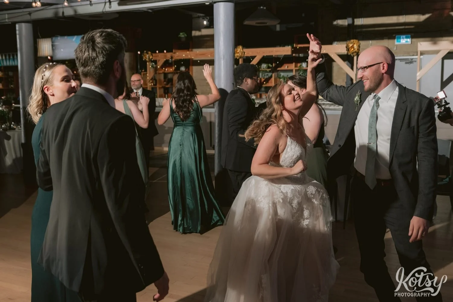 A bride and a white wedding gown and groom in gray shirt and green tie danced with big smiles and a crowd of people during their Second Floor Events wedding reception in Toronto, Canada.